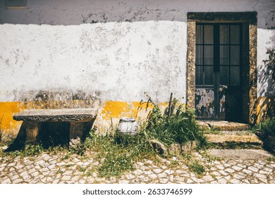 Weathered rustic wall with peeling white and yellow paint, old stone bench, ceramic jar, and overgrown grass next to a vintage door, ample copy space area on the left side