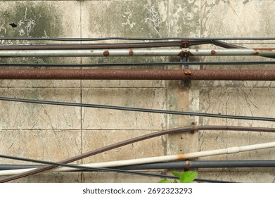 Weathered rusty electrical pipe installed on scratched and faded cinder block wall amid other pipes and cables in garden  
