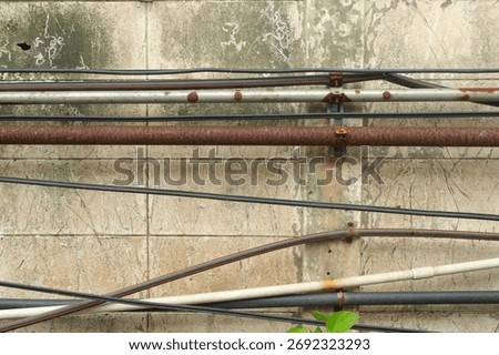 Weathered rusty electrical pipe installed on scratched and faded cinder block wall amid other pipes and cables in garden  