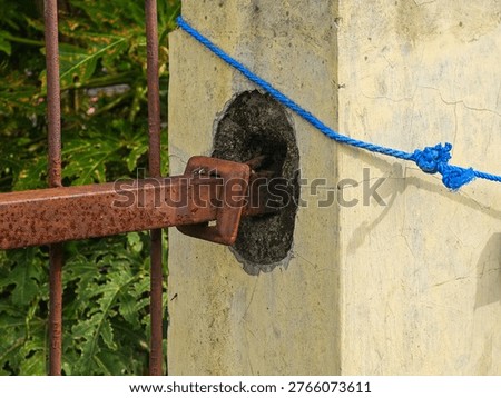 A weathered rusty gate latch secured to a cracked concrete pillar with a blue rope tied around the hole in the wall surrounded by green foliage.
