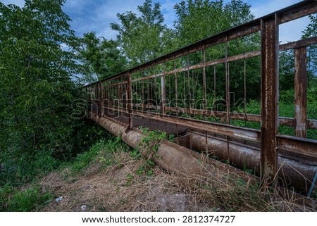 A weathered, rusty metal pedestrian bridge spans large industrial pipes surrounded by dense wild vegetation, captured from a low, wide-angle perspective at dusk. Ideal for urban exploration, industrial decay, and abandoned infrastructure themes.