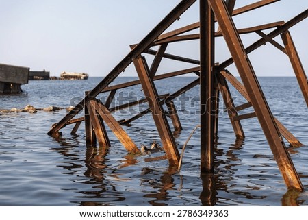 A weathered rusty metal structure extends into blue coastal waters beside a rocky shoreline. Broken bridge located at the Battery bay, Russia