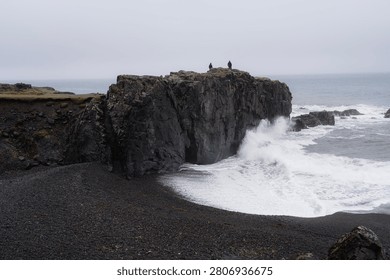 A weathered sea stack rises from a black sand beach on Iceland's east coast, hikers perched on top as powerful Atlantic waves crash below under a moody spring sky.