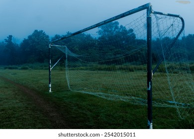 A weathered soccer goal stands alone on a misty field in the early morning, with the netting gently hanging, creating a tranquil yet melancholic scene of solitude and stillness. - Powered by Shutterstock