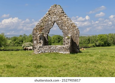 The weathered stone arch of Kilmacduagh Abbey, located in County Galway, Ireland, is captured against a backdrop of green fields and distant hills - Powered by Shutterstock