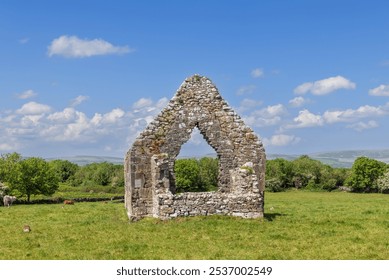 A weathered stone arch, part of the ruins at Kilmacduagh Abbey in County Galway, Ireland, stands alone in a green field. The ancient structure is framed by a clear blue sky and distant hills - Powered by Shutterstock