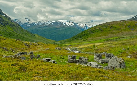 Weathered stone foundations of a remote mountain farmstead set within a lush green valley beneath dramatic snowy peaks and cloudy skies