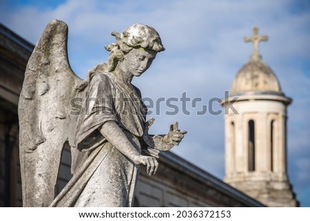 A weathered stone statue of praying angel on a grave with a bell tower in the background in Brompton Cemetery in London