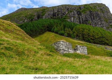 A weathered stone structure rests quietly in lush grass, surrounded by towering mountains and trees, evoking a sense of history and nature. - Powered by Shutterstock