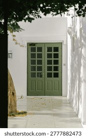 A weathered tree trunk frames a pair of classic olive-green double doors with glass panes, set against a bright white stucco wall casting dappled shadows from overhead foliage, evoking a serene Mediterranean atmosphere.