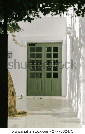 A weathered tree trunk frames a pair of classic olive-green double doors with glass panes, set against a bright white stucco wall casting dappled shadows from overhead foliage, evoking a serene Mediterranean atmosphere.