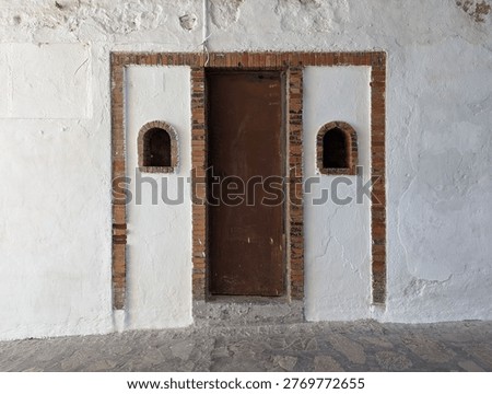 Weathered white-plastered wall with a central rustic brown metal door framed by an exposed red brick surround, flanked by two symmetrical arched brick niches set into the plaster, above a rough cobblestone floor, evoking the timeworn historic colonial charm