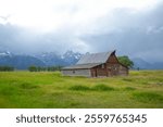 Weathered wooden barn sits in a grassy field with the stunning Teton Mountain range under a cloudy sky