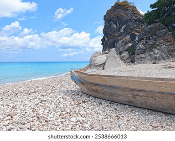 A weathered wooden fishing boat rests on a pebbled shore by a clear turquoise sea, with a rugged rock formation and vibrant blue sky, embodying coastal serenity - Powered by Shutterstock