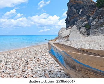 A weathered wooden fishing boat rests on a pebbled shore by a clear turquoise sea, with a rugged rock formation and vibrant blue sky, embodying coastal serenity - Powered by Shutterstock