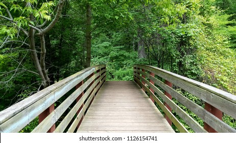 Weathered wooden footbridge & railings with rusted iron vertical supports, partially shaded by trees & foliage in the distance. Perspective view. Captured on a tranquil summer afternoon.