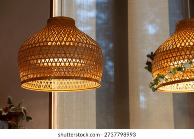 weaving bamboo lantern with A string of lights wire bulbs. Two lamps with a woven design are lit up and hanging from the ceiling. The lamps are placed in front of a window, which is partially covered by a curtain