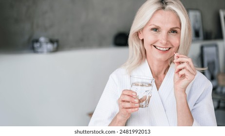 Web banner view of happy mature woman holding glass of still water, and vitamin pill with calcium and collagen. Mid age female feel good, smiling nice, taking dose of daily multivitamins at home - Powered by Shutterstock