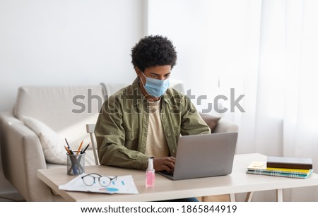 Web based learning during covid-19 pandemic. Teenage male student in medical mask attending online class on laptop at home. Black youth communicating with teacher or fellow students on webcam