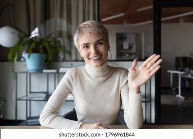 Web camera view smiling young short haired businesswoman sitting at table, waving hello gesture, welcoming client at professional online consultation or holding video call with partners at office.