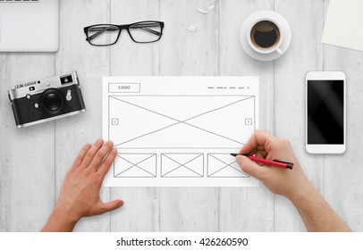 Web Designer Draw Layout Of Website On Paper. Top View Of Work Desk With Computer, Phone, Camera, Glasses, Coffee.