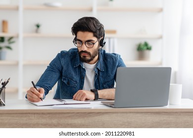 Web-based education concept. Arab man in headphones writing in notebook during online lecture on laptop, sitting at home interior. Concentrated guy doing homework and listening to webinar