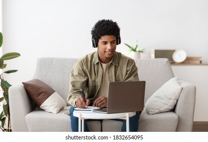 Web-based Education Concept. Happy Teen Guy In Headset Taking Notes While Communicating Online With Tutor On Laptop. Funky African American Adolescent Making Homework For His Online Course