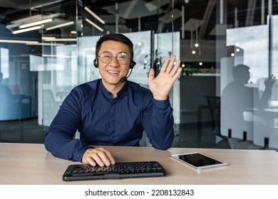 Webcam view, portrait of successful tech support and customer service worker, man smiling and looking at camera waving greeting hello, asian working inside modern office building