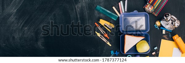 website header\
of lunch box with apple and sandwiches near school stationery on\
black chalkboard, top\
view
