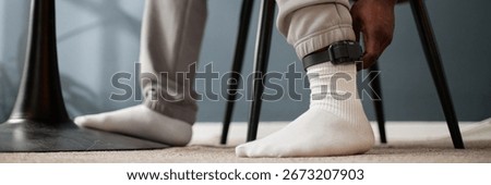 Website header shot of Black man wearing electronic ankle monitor sitting indoors adjusting device on ankle during house arrest, lower legs and hand visible, focus on equipment and subject action