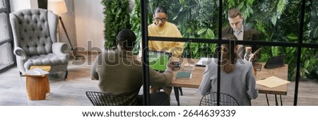 Website header shot of diverse group of young adult and middle aged professionals collaborating around table in modern office with plant wall, while using digital tablet and working with documents
