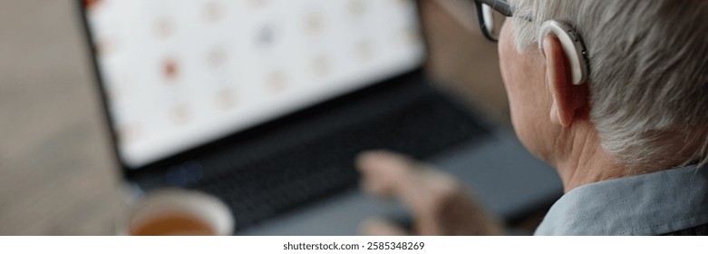 Website header shot of elderly man wearing hearing aid behind ear using laptop at home with help of assistive technology, copy space