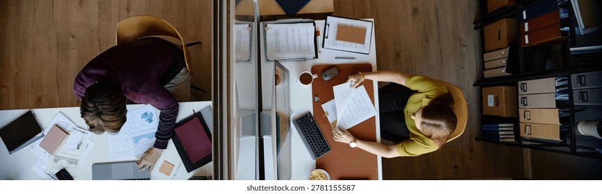 Website header shot of middle aged business people working at separate desks, reviewing documents and laptop data across office divider, overhead view. Ideal for teamwork, finance, productivity themes