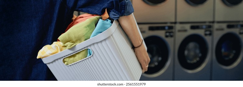 Website header shot of unrecognizable woman carrying basket with dirty laundry preparing colored clothes for washing in coin operated laundromat, copy space - Powered by Shutterstock