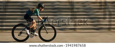 Website header of Side view of professional female cyclist in cycling garment and protective gear riding bicycle in city, rushing and passing buildings while training outdoors on a daytime