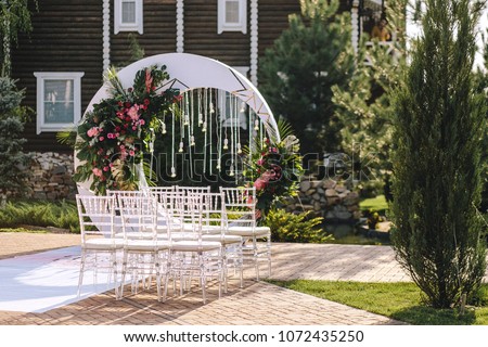 Wedding arch in the form of a white moon with floral compositions in a tropical style with leaves of a monster decorated with light bulbs. Transparent chairs for a ceremony in the street in the summer
