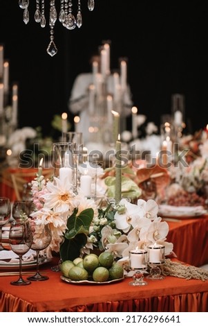 Wedding banquet table decor with orange tablecloth. In the foreground are lime fruits on a tray, white candles in candlesticks, black glasses. In the background are white flowers, candles, green leave