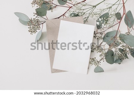 Wedding, birthday stationery composition. Blank greeting card, invitation mockup and craft envelope. Green Eucalyptus populus branches, Gypsophila flowers isolated on table background. Flat lay, top.