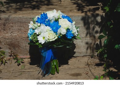 Wedding Bouquet Of Daisies, White Roses, Blue Chrysanthemums And Green Leaves Wrapped In Blue Ribbon On A Sunny Summer Day On A Background Of Wooden Stairs. Bride's Wedding Accessory On A Happy Day.