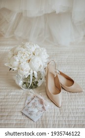 Wedding bouquet of peonies flowers in a vase stands on the bed of the newlyweds with invitations and shoes on the background of the bride dresses