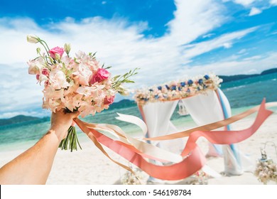 wedding bouquet of pink and white flowers at arm's length, on a background of an arch of white sand beach paradise island.
Wedding ceremony on a tropical island.