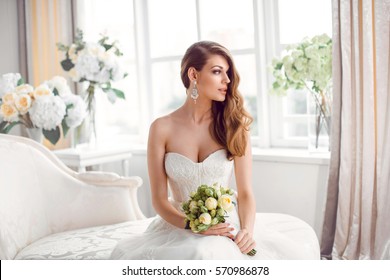 Wedding. Bride in beautiful dress sitting on sofa indoors in white studio interior like at home. Trendy wedding style shot. Young attractive caucasian brunette model like a bride against big window