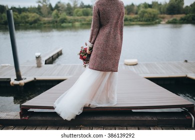 Wedding. The bride's bouquet. Wedding bouquet . The bride in a white dress standing on a pier at the lake and holding a bouquet of red and pink flowers with berries and greens with a marsala ribbon