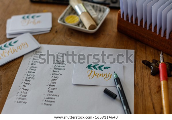 Wedding calligraphy - guest list and\
handwritten place cards with names. Golden writing with green\
flourish on white paper. Gold paint, nib pen, card\
holder.