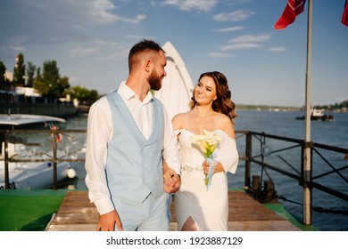 A Wedding Ceremony By The Water On The Dock. Romantic Wedding In The Sea Style In A Yacht Club. Beautiful Bride With Long Curls In An Elegant White Dress And The Groom In The Blue Suit.