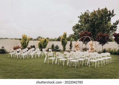Wedding Ceremony In The Garden For The Bride And Groom. The Arch Is Made Of Fresh Flowers And Dead Wood, Reeds. Outdoor Wedding Ceremony. Wooden Chairs For Guests