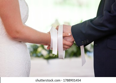 Wedding ceremony moment, bride and groom's hands tied with white ribbons, white wedding dress, dark suit