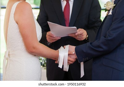 Wedding ceremony moment, bride and groom's hands tied with white ribbons, white dress, dark suit , wedding celebrant's hands holding paper