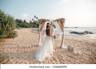 Wedding ceremony on a tropical beach. Happy groom and beautiful bride kissing under the arch decorated with flowers on tropical sand beach wih palms. Sea on the background