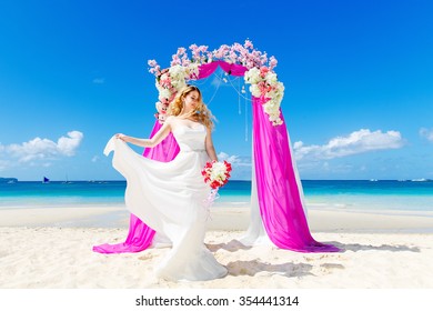 Wedding ceremony on a tropical beach in purple. Happy blond bride with wedding bouquet under the arch decorated with flowers on the tropical sand beach. Wedding and honeymoon concept.